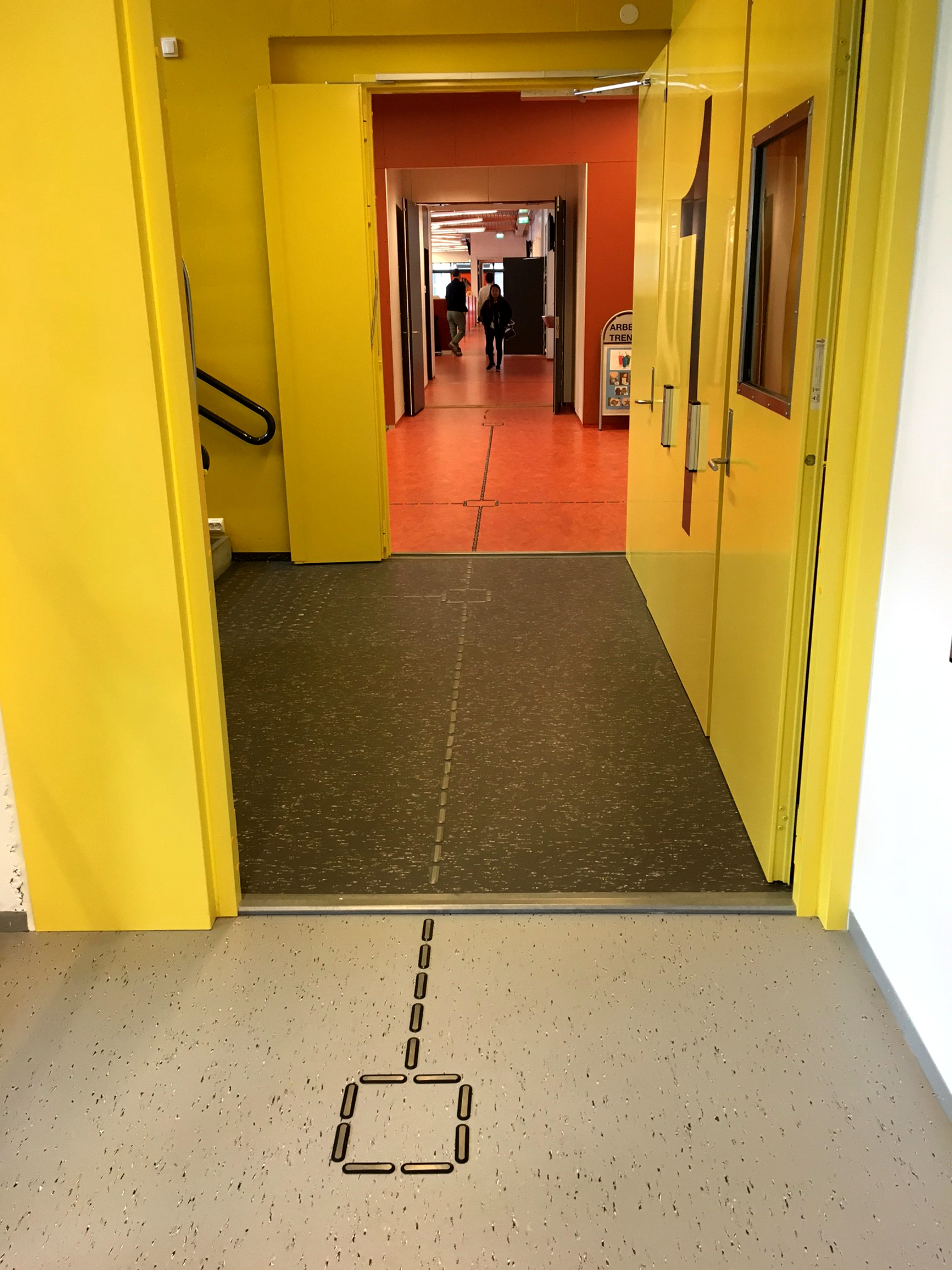 FARGESONER: Fargede soner veileder. Korridoren nærmest har grått gulv med hvite spetter, mens bi-trappeløpet har samme belegg i en mørkere farge mot gule vegger og dører. En rød allmenning, en frisone, fører inn til kantineområdet med samme gulv. 