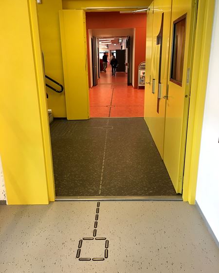FARGESONER: Fargede soner veileder. Korridoren nærmest har grått gulv med hvite spetter, mens bi-trappeløpet har samme belegg i en mørkere farge mot gule vegger og dører. En rød allmenning, en frisone, fører inn til kantineområdet med samme gulv. 