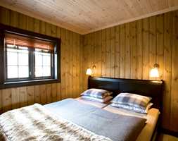 Soverom med lys panellakk i tak og interiørbeis på vegg. Vinduene i den grå farge, sengen er i sort og sengeutstyr i matchende blå- og bruntoner.