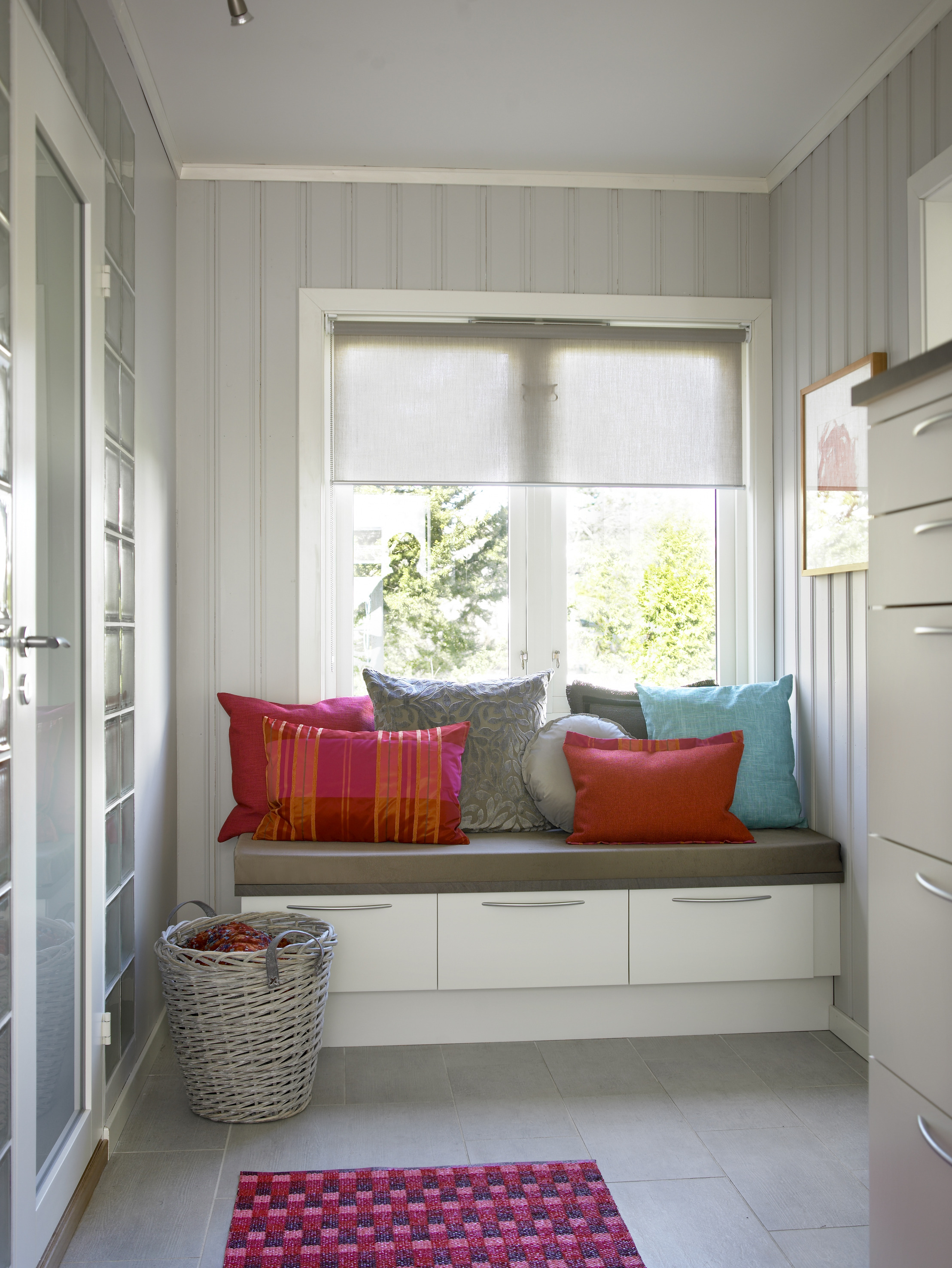 Sofabenken er gjort til et blikkfang i innredningen. Den inviterer til å bruke rommet til mer enn en rask passasje. Putene er sydd i en dristig kombinasjon av farger og materialer, og tilfører fargeglede i den ellers så rolige rammen. 