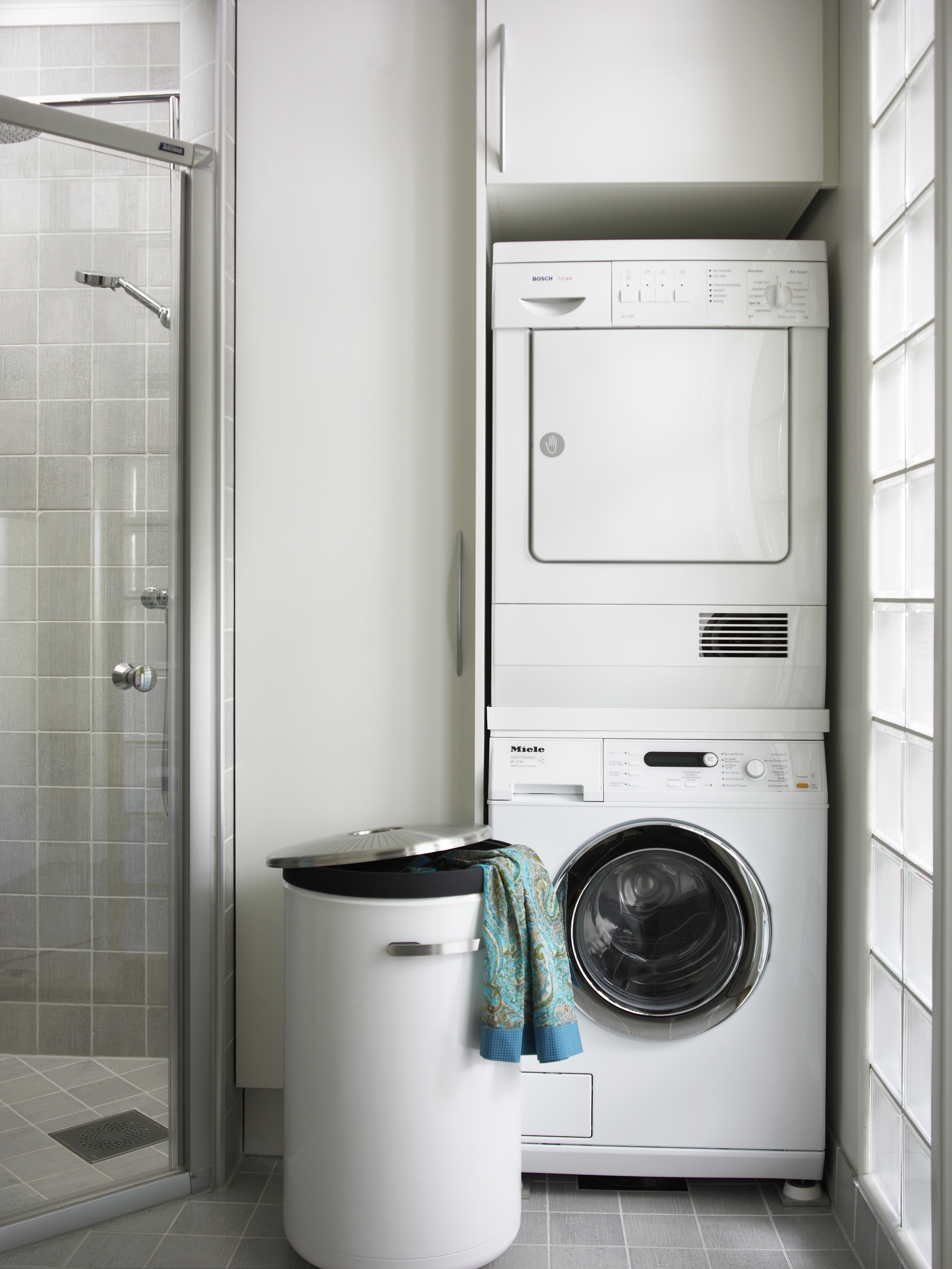 Vaskemaskin og tørketrommel over hverandre sparer plass. Et høyskap skjuler vaskeremedier. Glassbyggesteinene tilfører naturlig lys fra entréen og øker romfølelsen.  