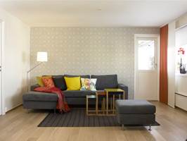 Den ene kortveggen i stuen er møblert med ny grå sofa med sjeselongdel og frittstående pall (Milano fra Bolia). Sofaen er fylt med puter i friske farger og mønster (Hilmers Hus). Det store lappeteppet er sydd sammen av ulike mønster i silke. Veggen bak sofaen er tapetsert med en retro tapet i gråtoner (Ecco Fond 05 5922 fra Borge). Settbordene (Nesting tables) er designet i 1926 av Josef Albers for Vitra. Bordene har ben av eik og med topplate av farget akrylglass.
