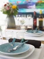 Bordet er dekket til fest. Turkis dekketallerkener på løpere i lysblå lin med brune servietter til (Hilmers Hus). Serviettringer av turkis perler og en vakker sommerbukett pynter opp.