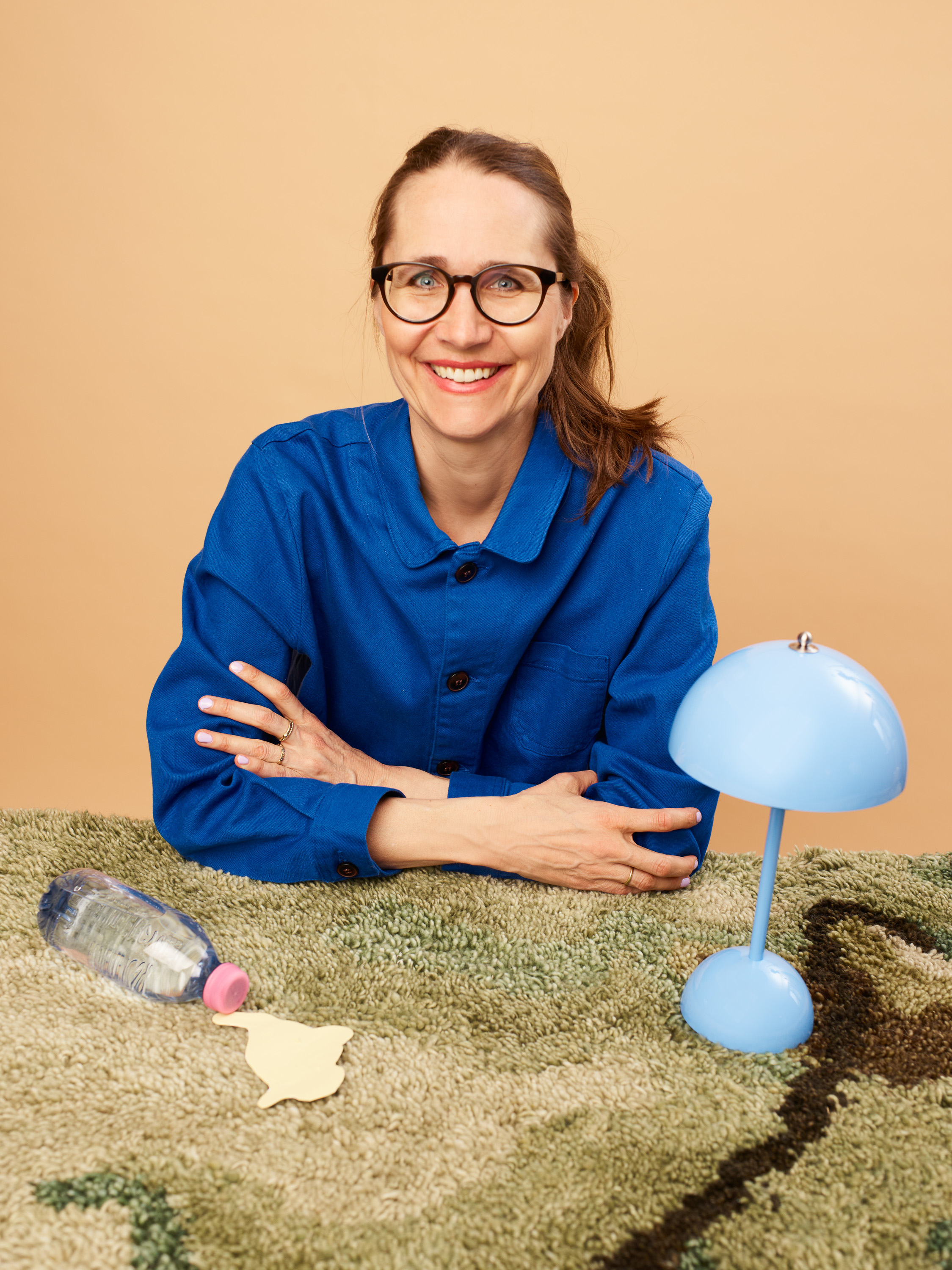 dame i blå bluse og briller sitter ved et bord med et grønt teppe der det står en blå lampe på